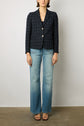 Woman wearing a dark plaid blazer, light blue wide-leg jeans, and black open-toe heels standing against a plain light-colored wall.