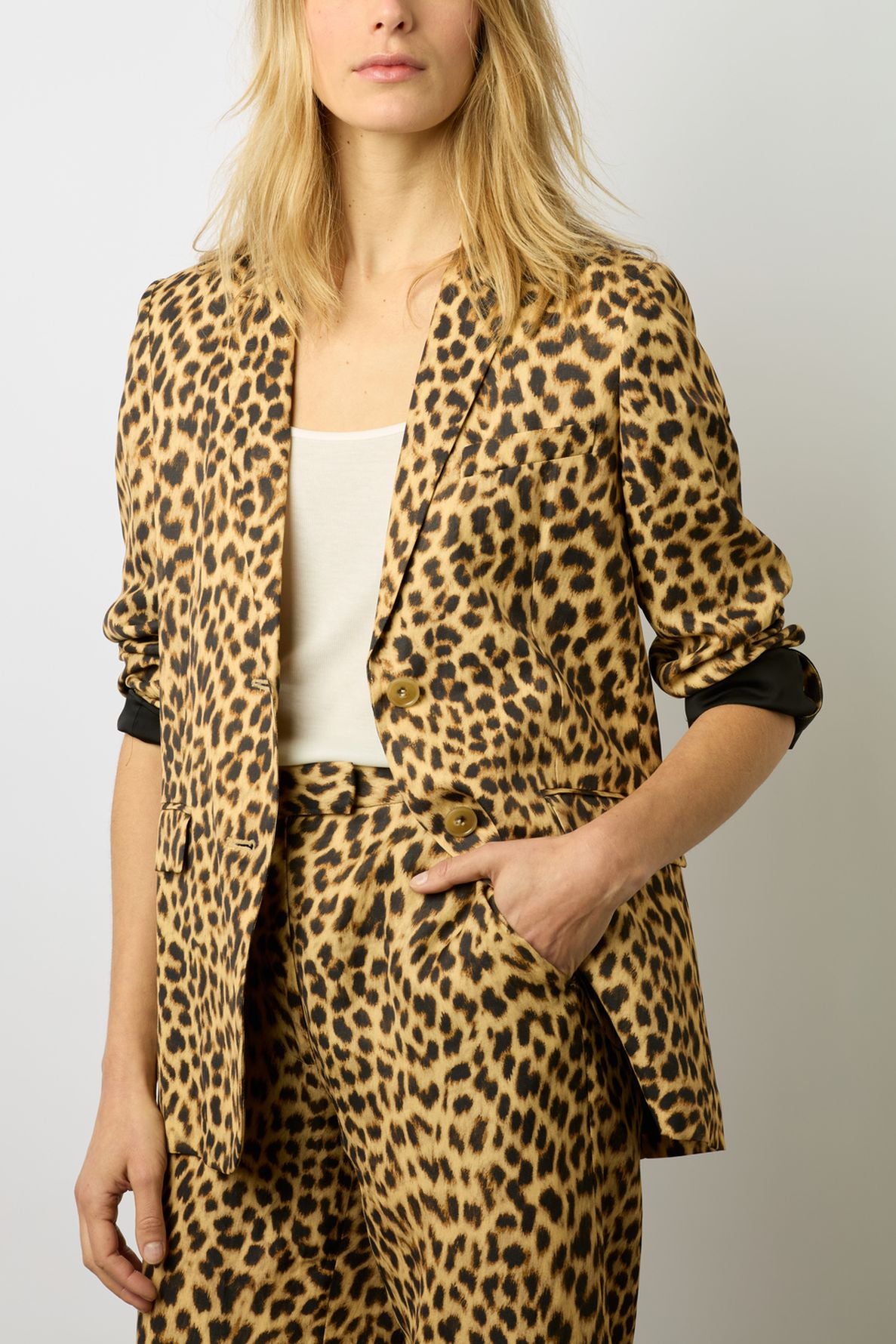 A woman wearing a veste de tailleur imprimé léopard with a white top underneath, standing with one hand in her pocket against a plain background.