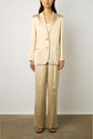 A person stands against a plain background wearing a shiny, light gold veste tailleur satinée with a blazer and wide-leg pants, paired with black open-toe heels. Their hands are relaxed at their sides.