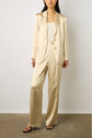 A woman stands against a white wall wearing a shiny beige satin suit with a matching blazer satiné and pants, a white top, and black sandals.