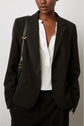 A person wearing a black blazer over a white button-up shirt, black pants, and carrying a black shoulder bag with a gold chain strap. The background is plain and light-colored.