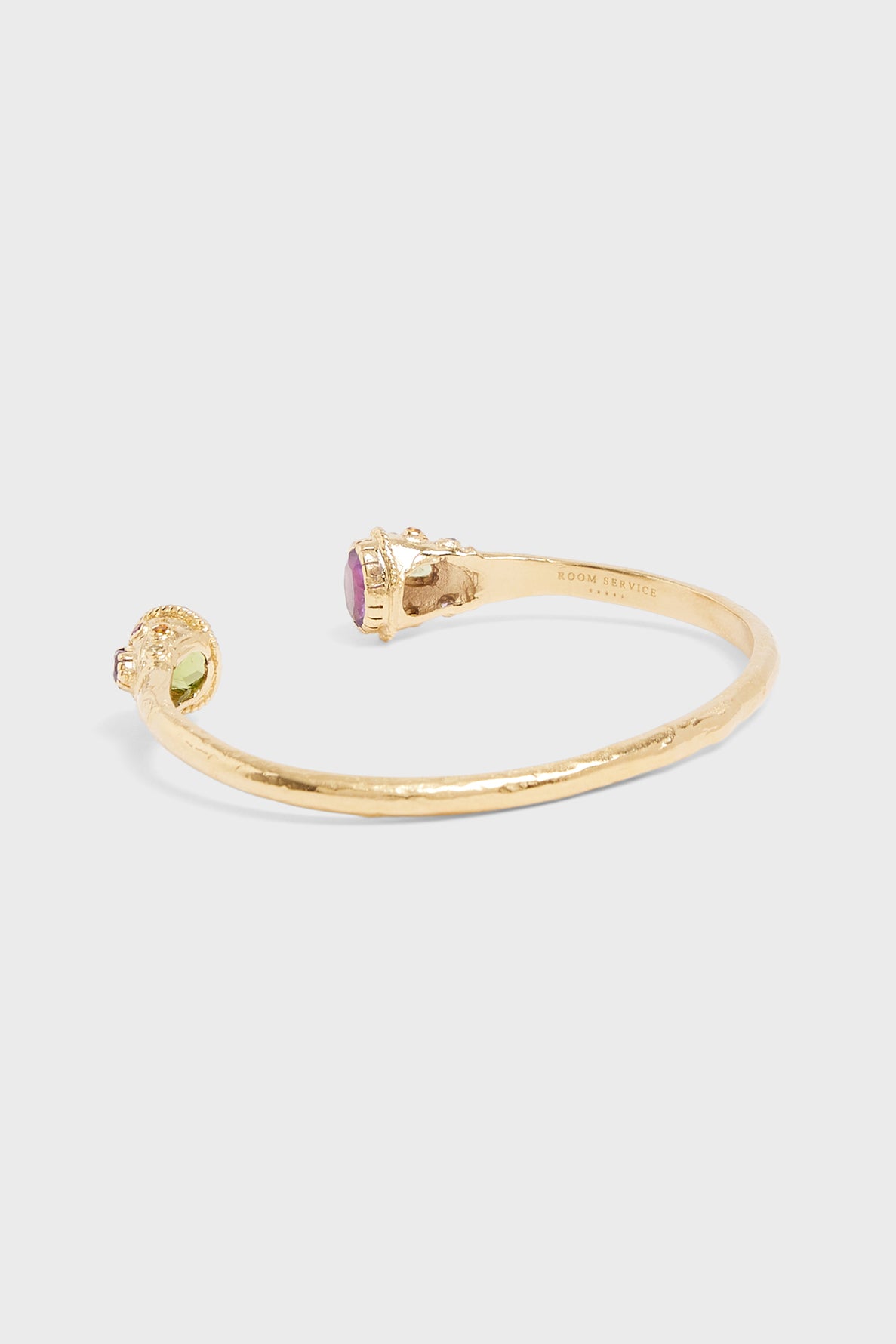 A gold open bangle bracelet laiton with a textured finish, featuring a green gemstone on one end and a pink gemstone on the other, displayed on a plain light background.