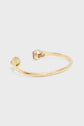 A gold open bangle bracelet laiton with a textured finish, featuring a green gemstone on one end and a pink gemstone on the other, displayed on a plain light background.