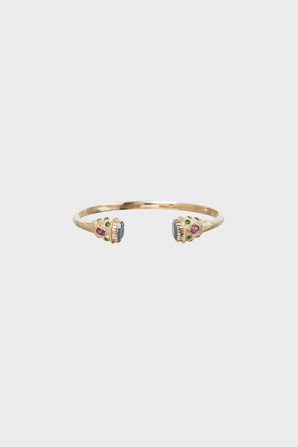 Thin gold cuff bracelet with two ornate ends, each featuring multicolored gemstones and blue central stones, displayed on a plain light gray background.