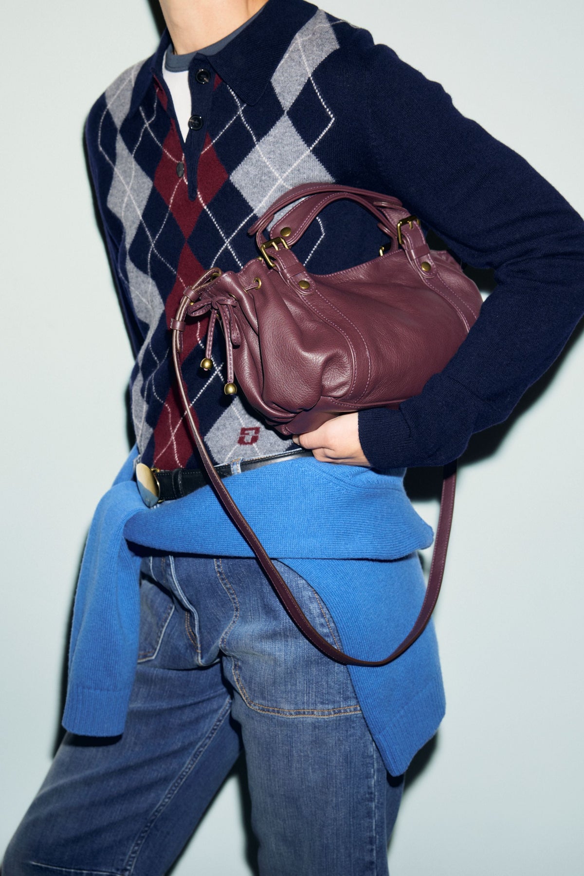 A person in an argyle sweater and jeans holds a burgundy sac bandoulière en cuir Micro 24. A blue sweater is tied around their waist. The background is plain and light-colored.