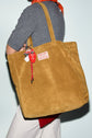 A person holds a large tan suede tote bag cuir with a red keychain and a patch that reads GERARD DAREL MEXICO PARIS. The person wears a gray top, white pants, and a red-orange scarf, embodying the effortlessly chic Gérard Darel style seen on the streets of Merci Paris.