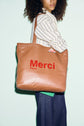 A person with curly hair holds a large tan Gérard Darel tote bag that says Merci Paris in red letters. They wear a striped shirt and blue jeans, standing against a pale background.